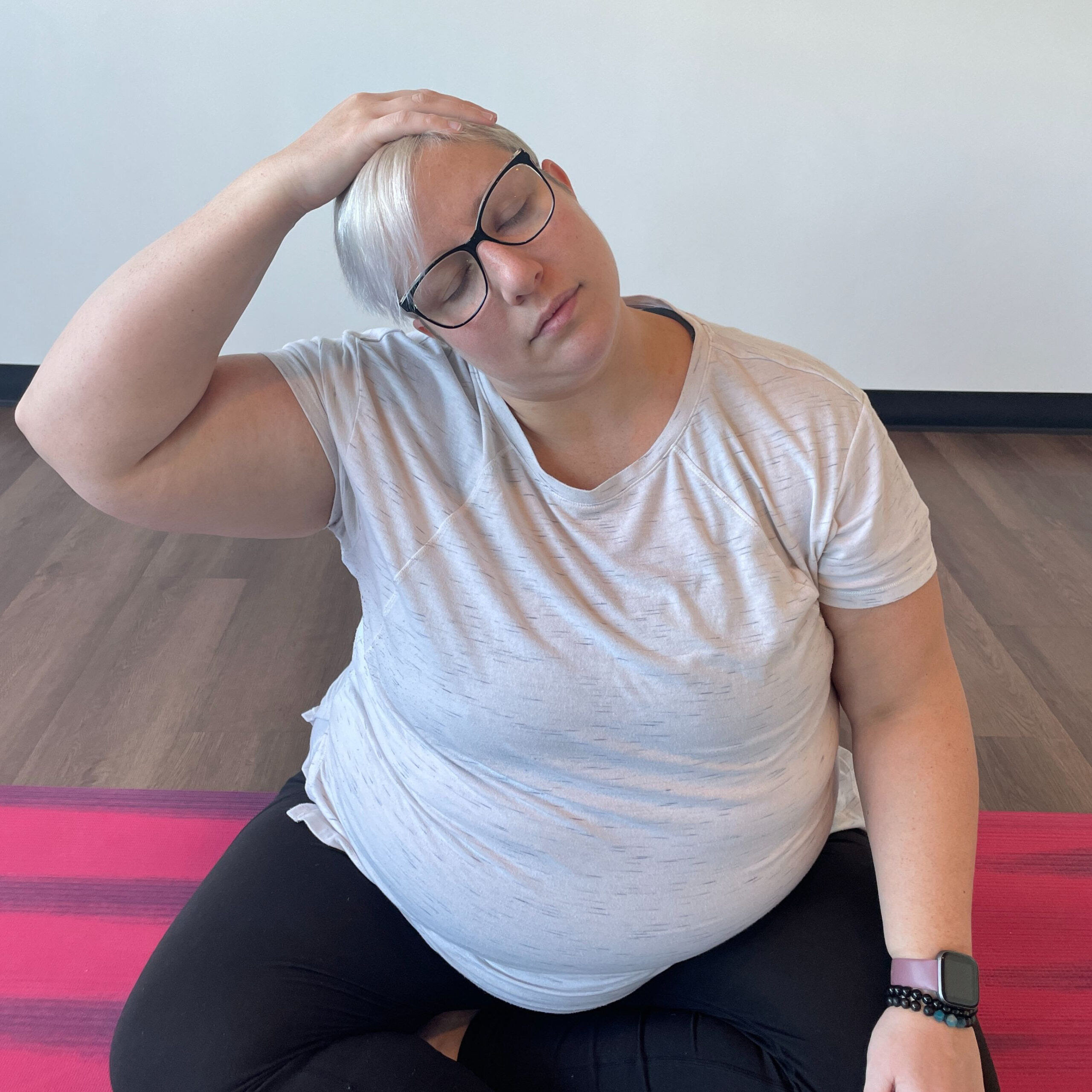 Karen Kirchman, a white woman with short blonde hair and glasses, sits on a pink yoga mat and practices stretching her neck.