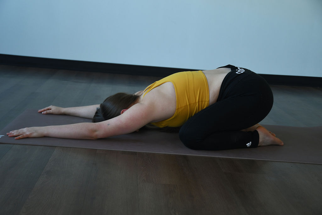 Zoe Lowden, a white woman with long brown hair, a yellow tank top, and black yoga pants, practices balasana on a tan yoga mat.