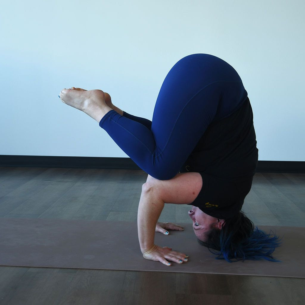 Kati Black, a white woman with dark blue hair, wearing a black shirt and dark blue yoga leggings, practices tripod headstand. 