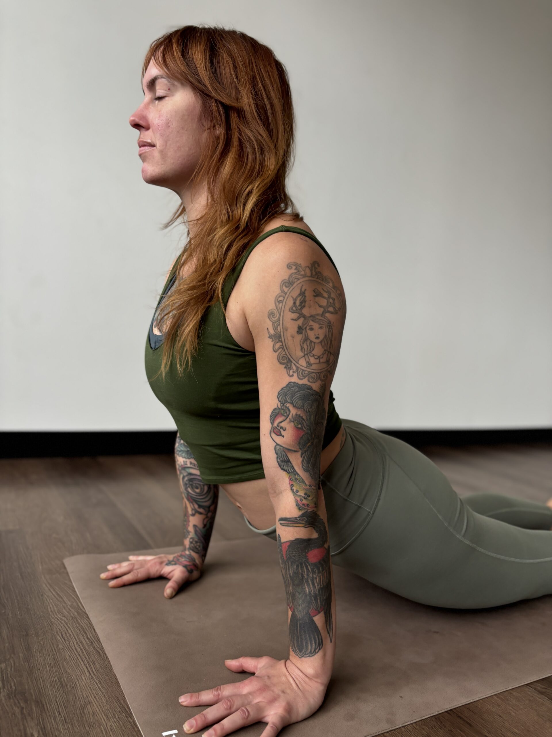 A white woman with red hair and tattoos wearing green leggings and a green tank top practices upward facing dog, a prone backbend, on a tan yoga mat. 