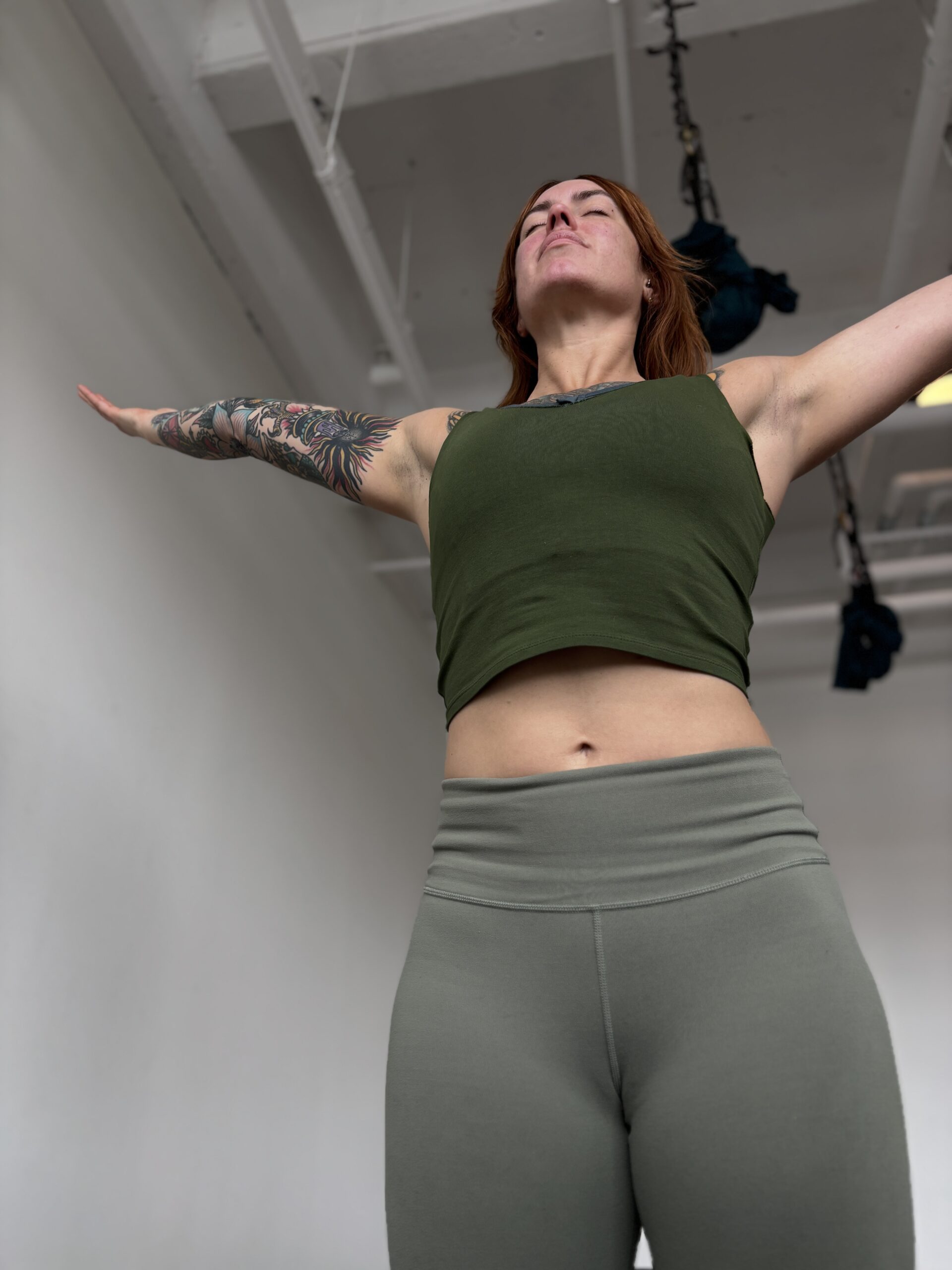 A white woman with red hair and tattoos practices urdhva hastasana reaching her arms overhead. She is wearing light green yoga pants and a dark green tank top.