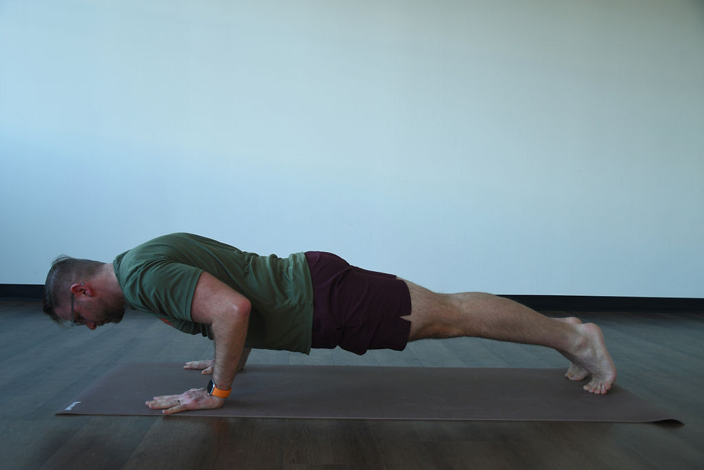 A white man with short hair and glasses, wearing a green shirt and black shorts practices chaturanga dandasana, a bottom of a pushup hold on a tan mat. 