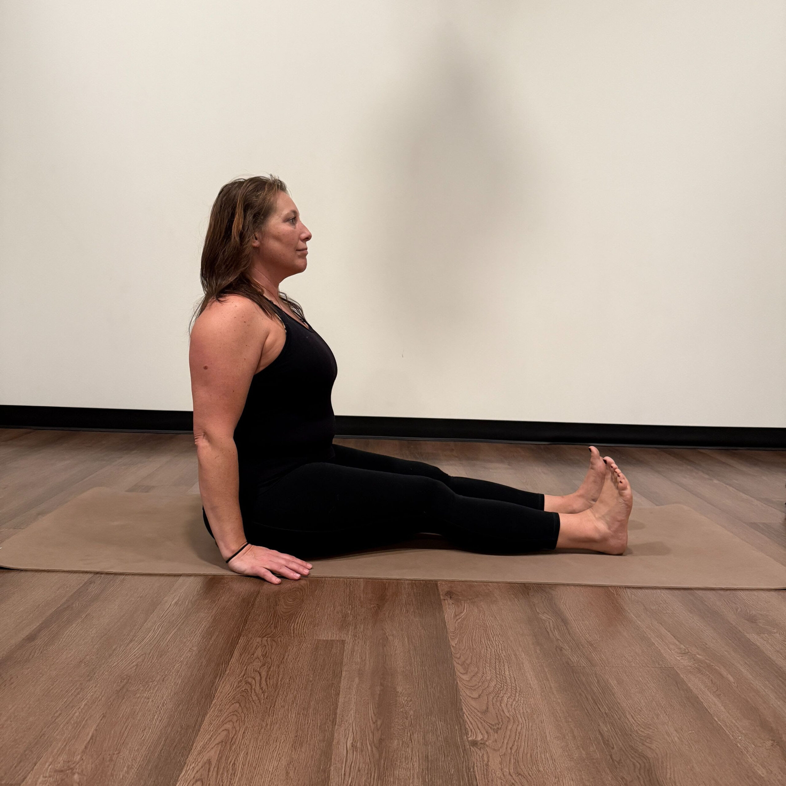 Kati Black, a white woman with brown shoulder length hair wearing a black tank top and black leggings practices dandasana on a tan yoga mat. 