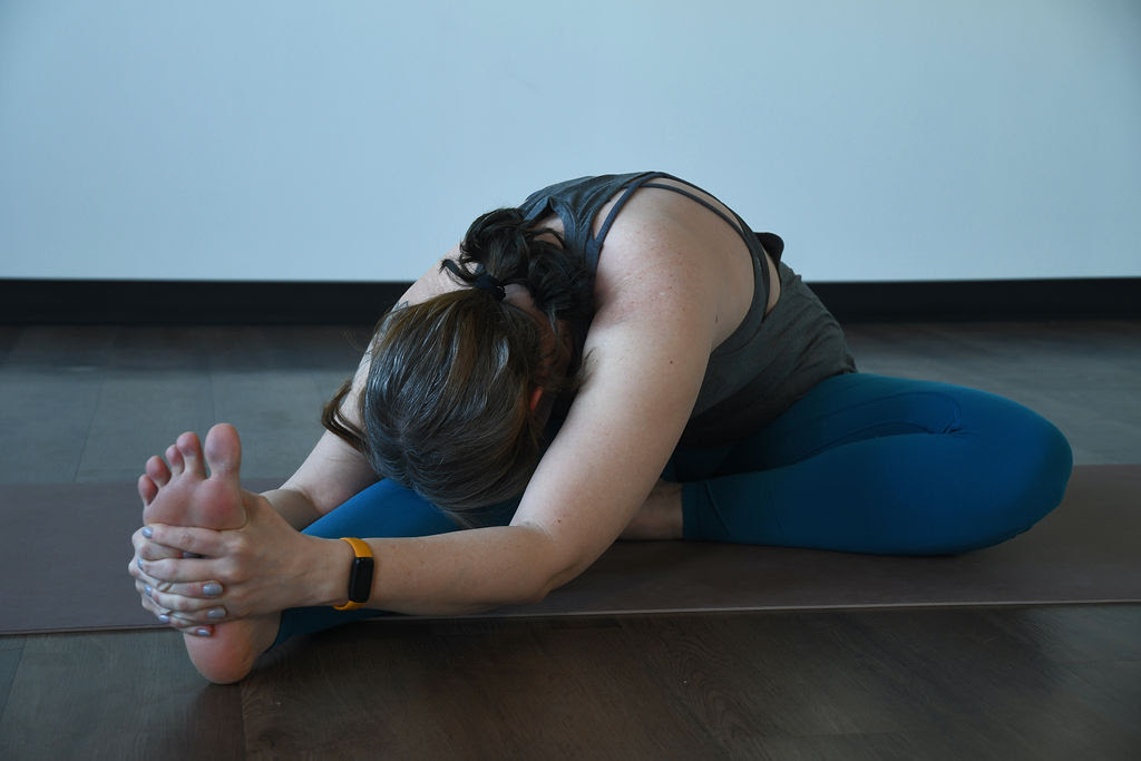 A white woman with brown hair in a ponytail wearing a grey tank top and blue leggings practices janusirsana, a seated stretch with one leg extended and the other leg in a bent position.