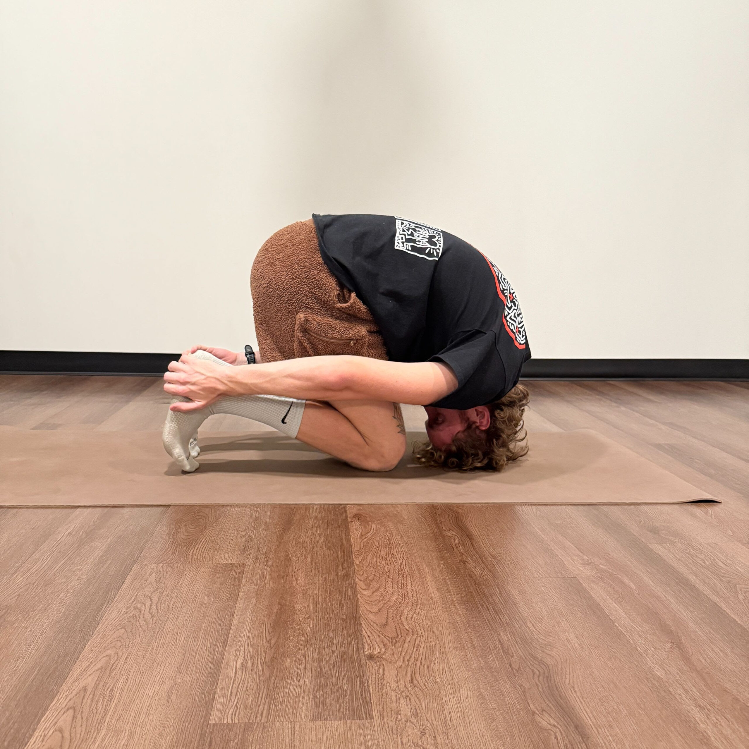 Jared France, a white man with curly tan hair practices rabbit pose on a tan mat. He is wearing fuzzy brown shorts, white socks, and a black shirt. 