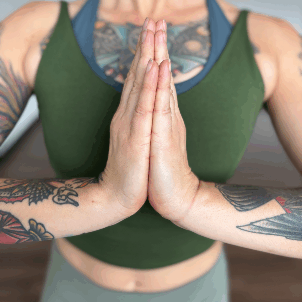 A white woman with tattoos, a green tank top, and light green leggings holds her hands together in anjali mudra.