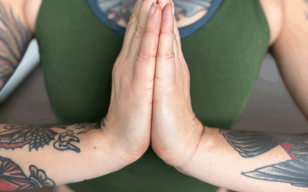 A white woman with tattoos, a green tank top, and light green leggings holds her hands together in anjali mudra.