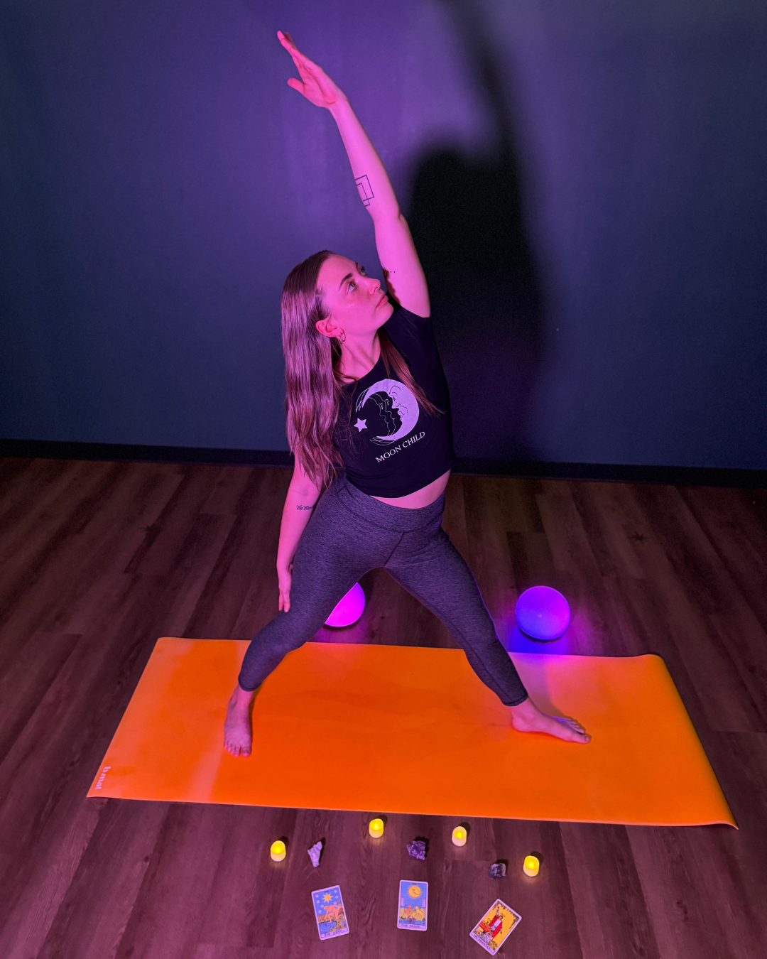 Zoe Lowden, a white woman with long brown hair wearing black leggings and a black crop top featuring the moon practices Sky Archer Pose on a yellow yoga mat in a dark room surrounded by candles and tarot cards.