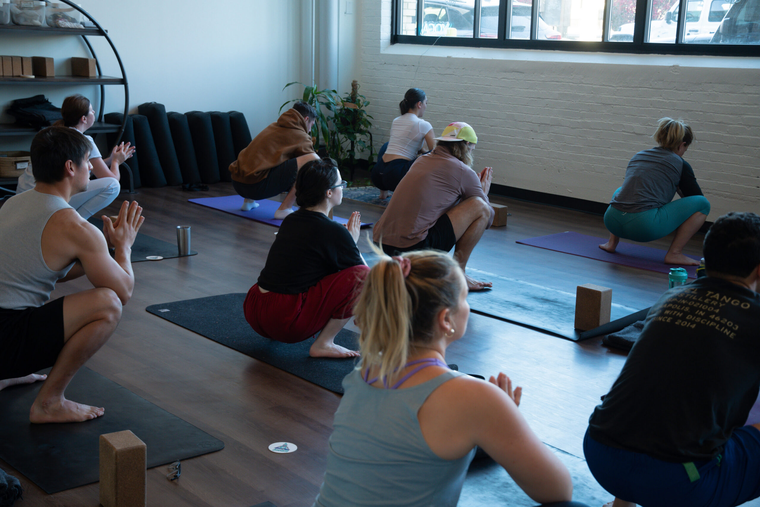 A group of yoga students sit in a well lit room practicing malasana pose. 