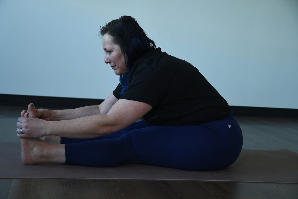 Kati Black, a white woman with dark blue hair, wearing dark blue leggings and a black short sleeve shirt practices paschimotanasana on a beige yoga mat