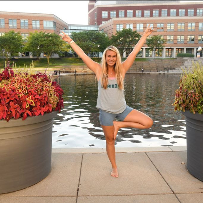 Celia Sadjadi, a white woman with blonde hair wearing light blue yoga shorts and a light blue tank top, practices tree pose while standing outside in front of a body of water. 