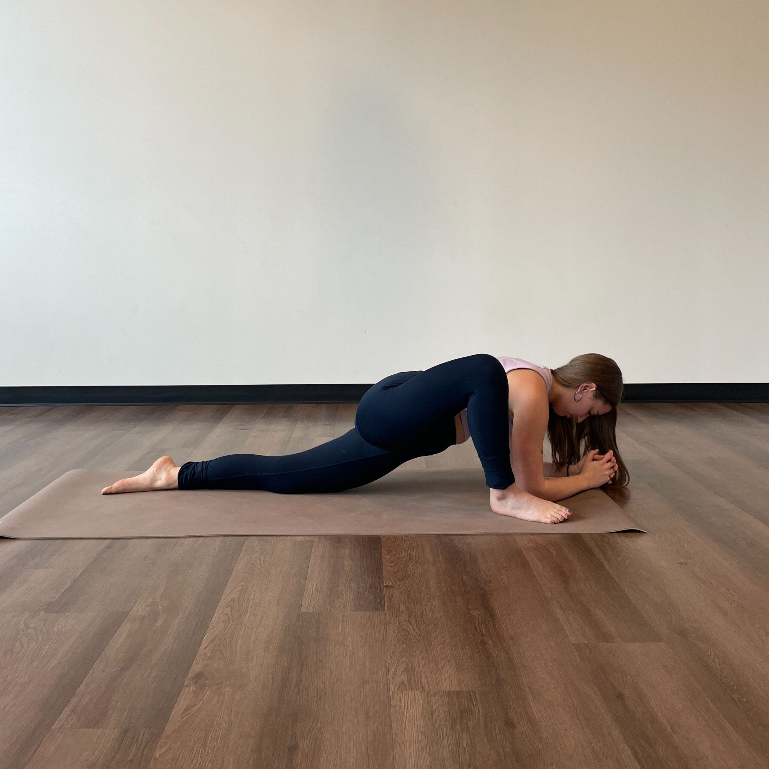 Zoe Lowden, a white woman with long brown hair wearing black leggings and a light purple tank top practices dragon pose on a beige yoga mat. 
