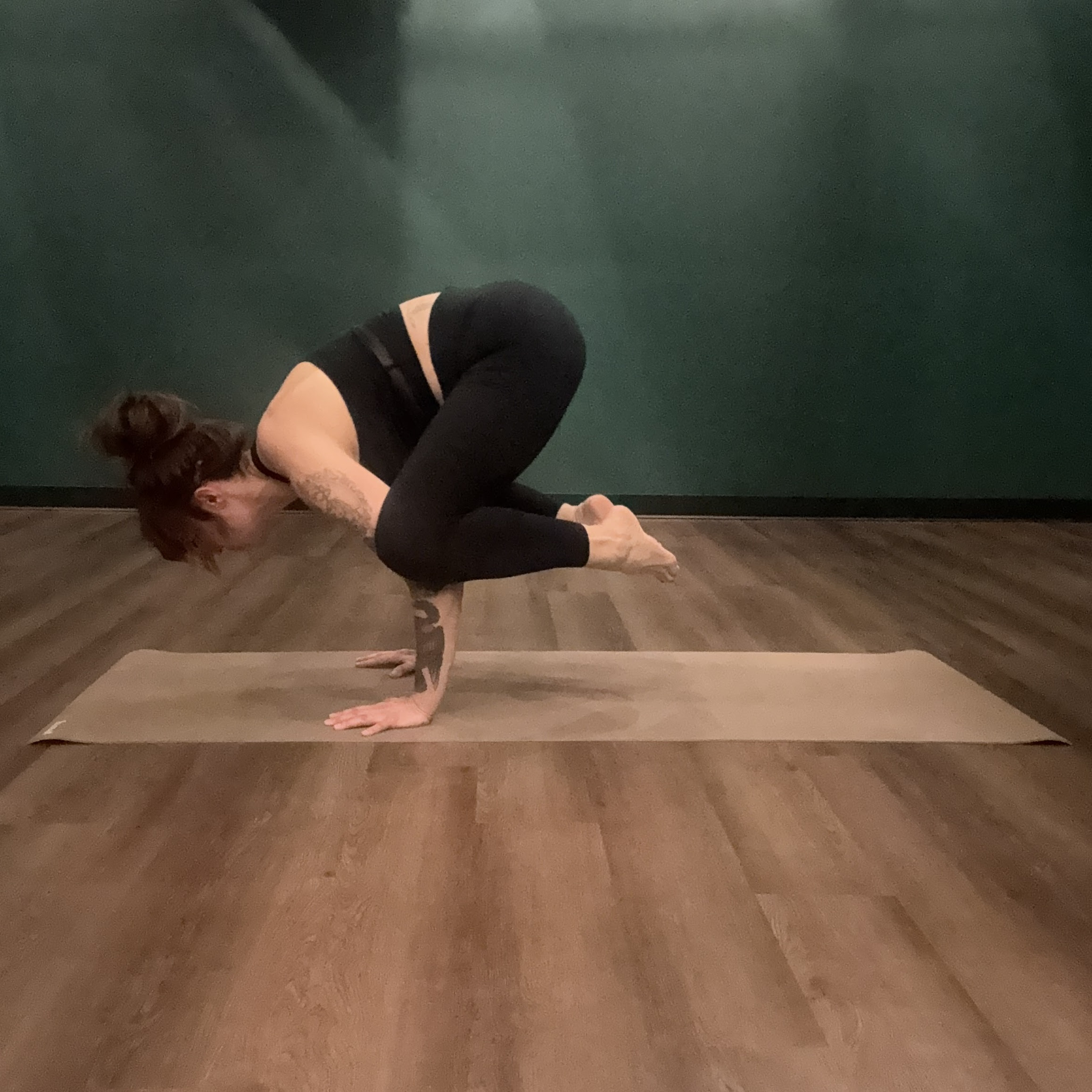 Marta Gruber, a white woman with dark red hair in a bun wearing black leggings and a black tank top practices crow pose on a beige yoga mat.