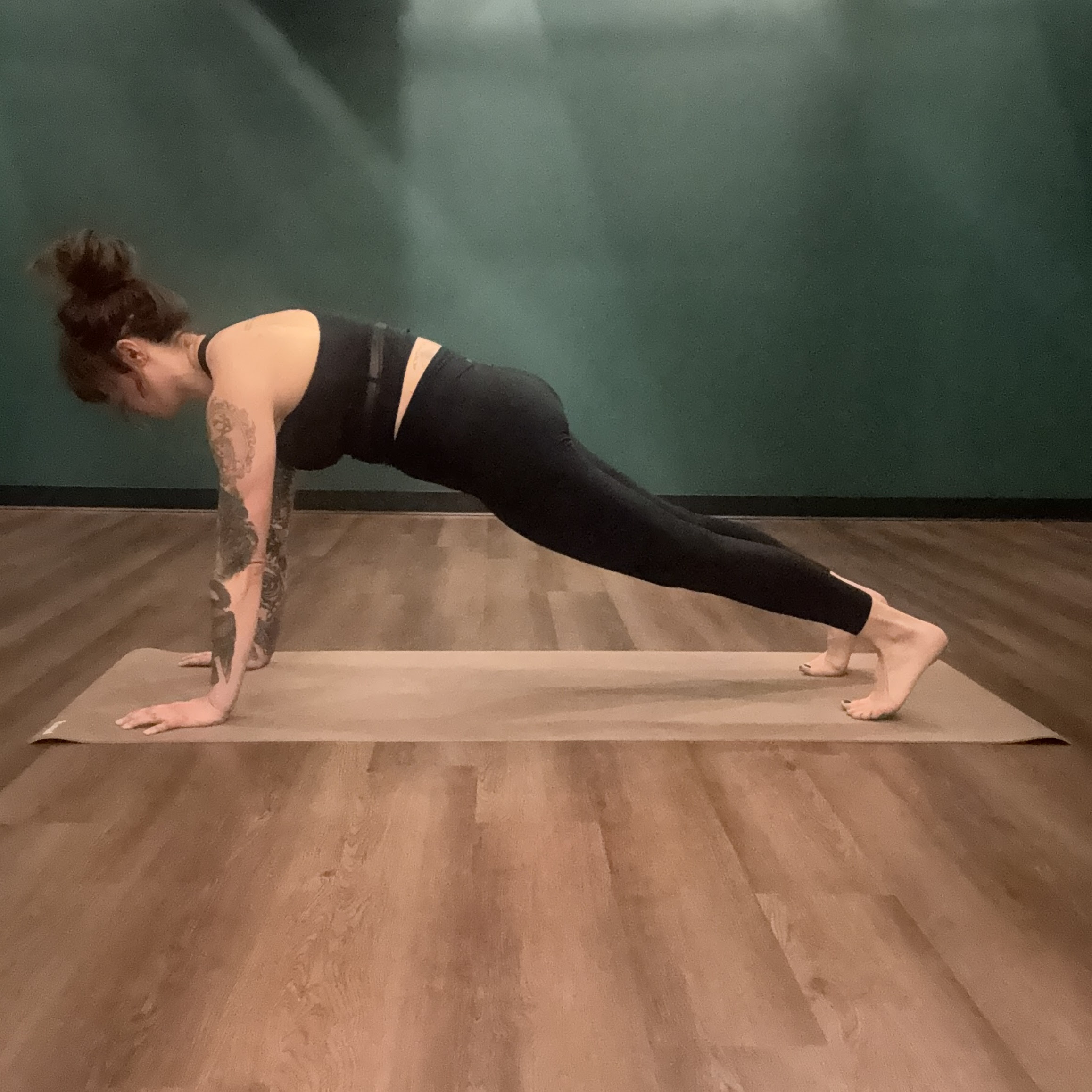 A white girl with red hair tied in a bun, wear a black tank top and black leggings, with tattoos on her arms, holds a plank pose position on a beige yoga mat. 