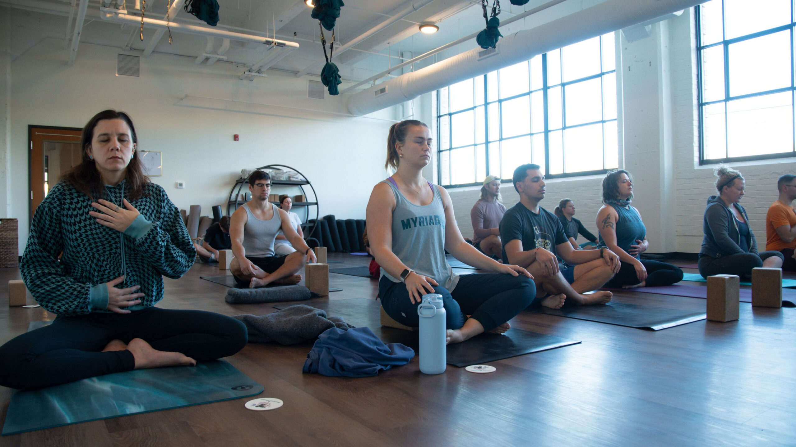 Students sit in a brightly lit yoga studio with white walls, large windows and wooden floors on yoga mats in sukhasana. 