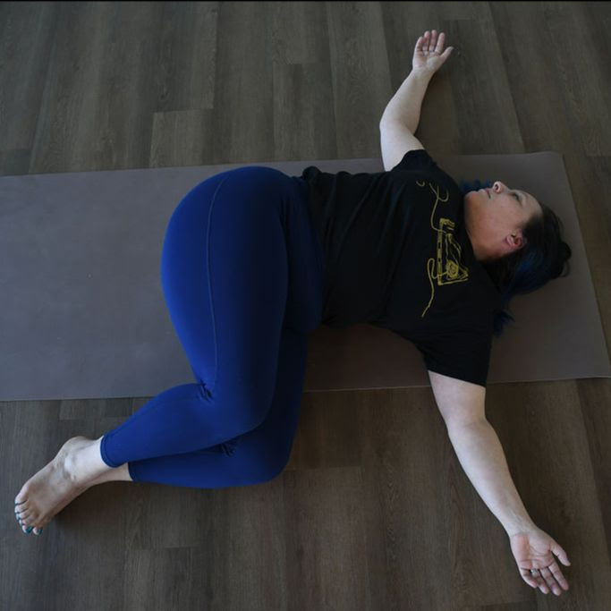 Kati Black a white woman with dark hair wearing dark blue leggings and a black tshirt practices supta matsyendrasana on a beige yoga mat.