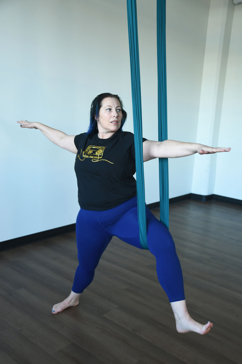Kati Black, a white woman with blue hair, blue leggings and a black shirt practices warrior II pose using an aerial silk as a prop.