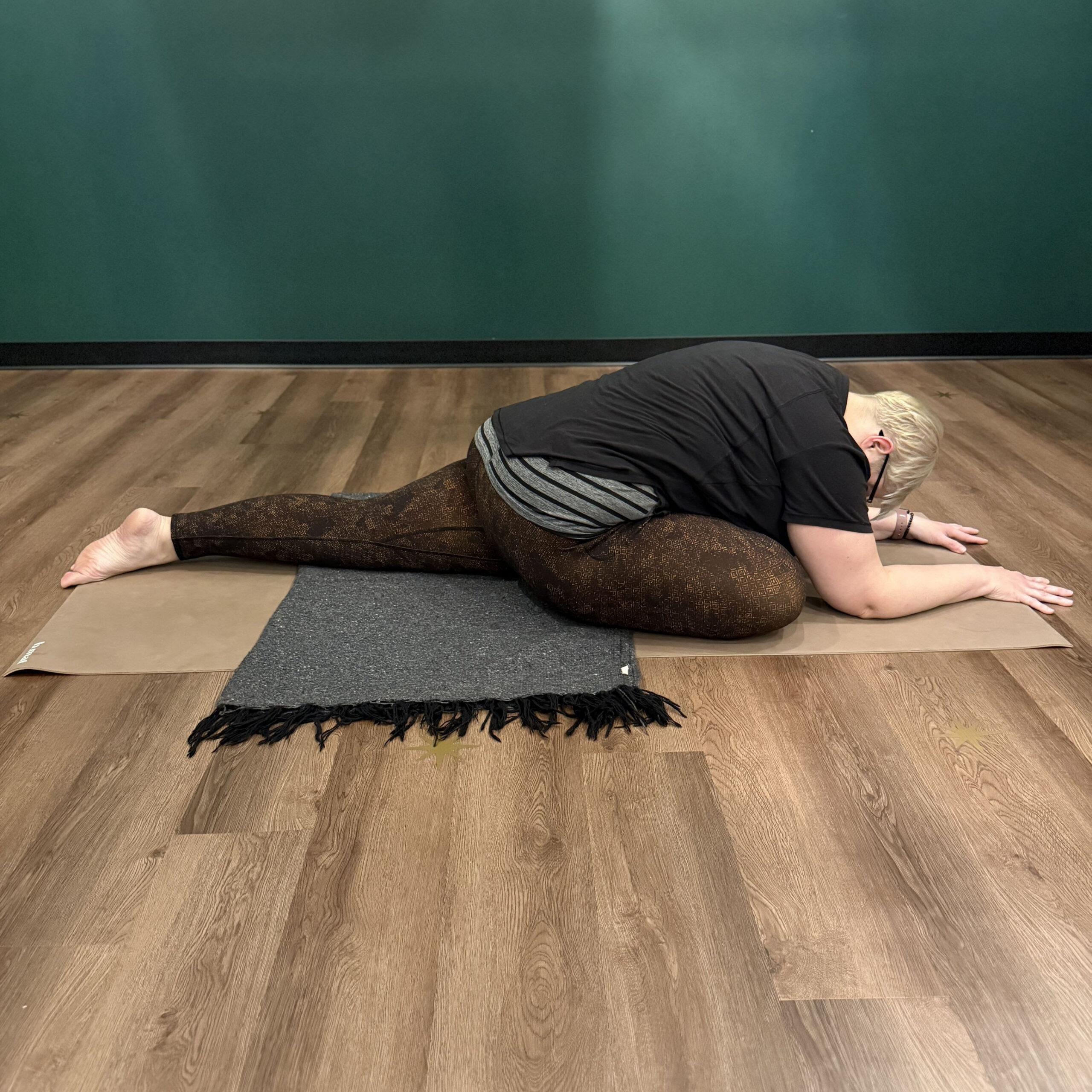 Karen Kirchman, a white woman with short platinum blonde hair wearing brown leggings and a black shirt practices kapotasana on a beige yoga mat with a blanket beneath her hips for support.