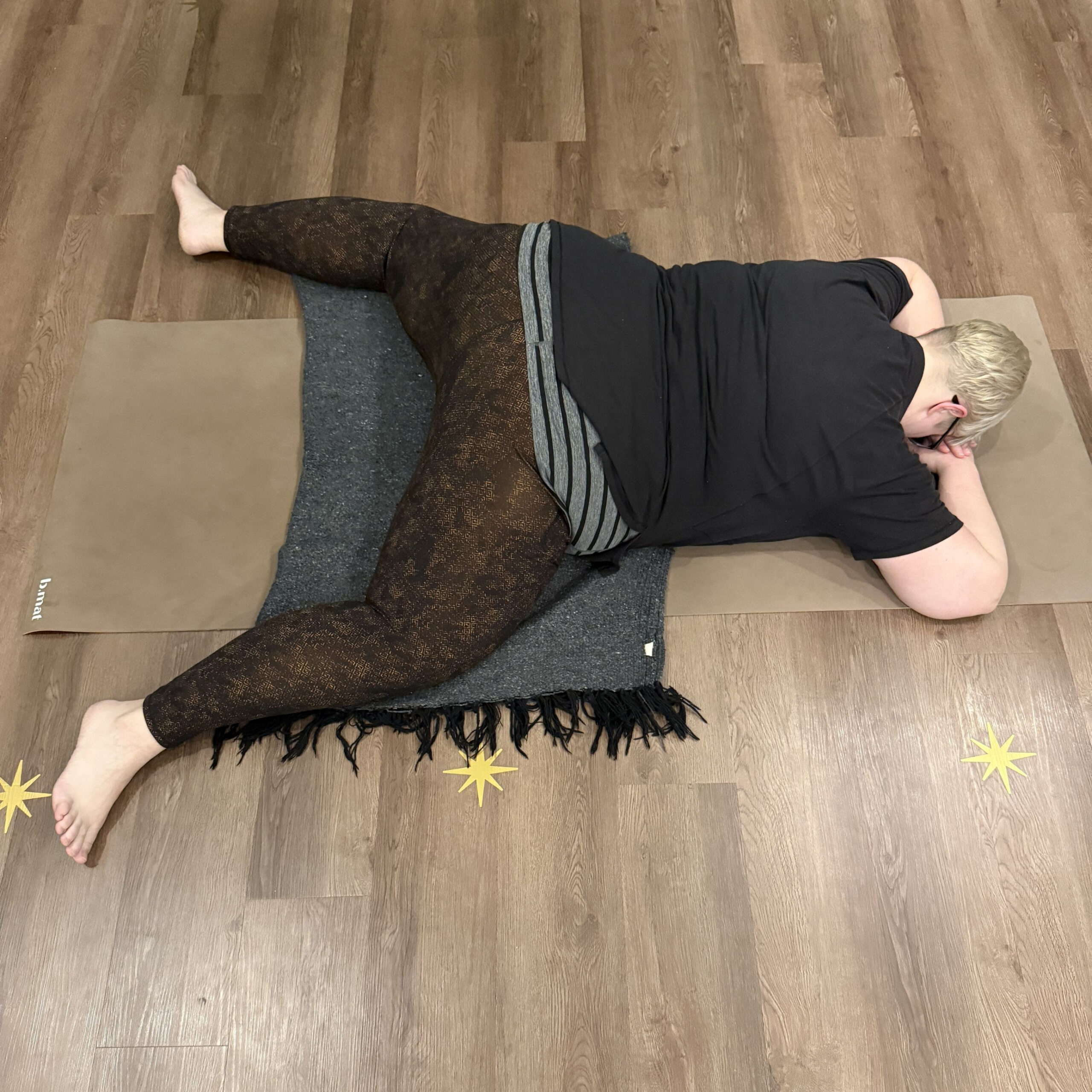 Karen Kirchman, a white woman with short platinum blonde hair wearing brown leggings and a black shirt practices mandukasana on a beige yoga mat using a blanket to pad her knees. 