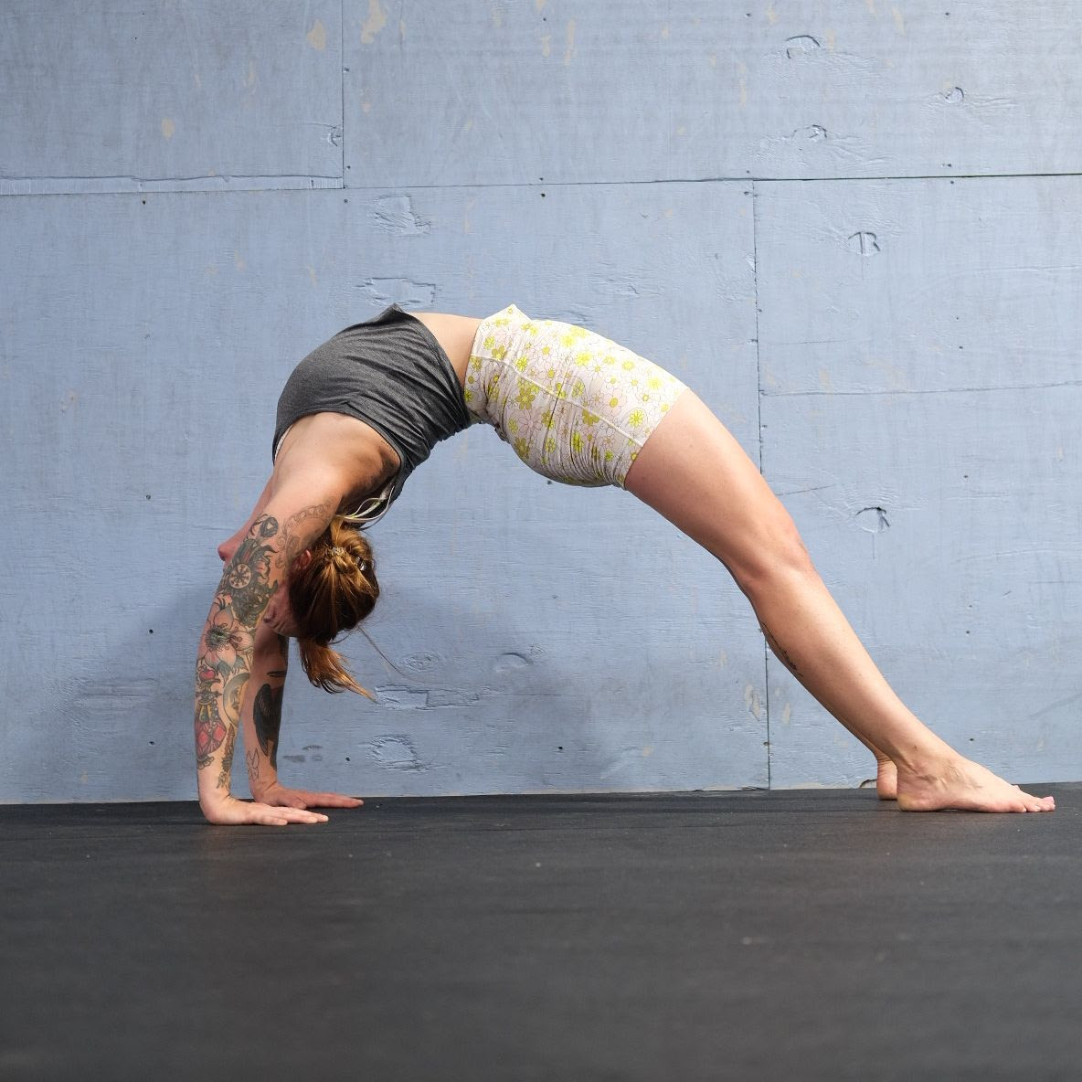 Marta Gruber, a white woman with red hair wearing white yoga shorts and a grey tank top practices urdhva dhanurasana. 