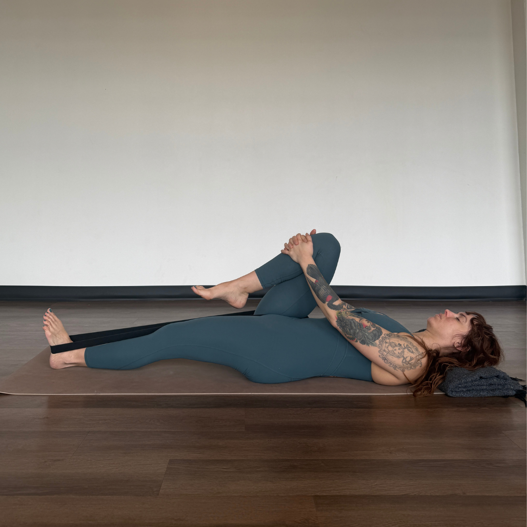 Marta Gruber, a white woman with red hair wearing a teal yoga jumpsuit lies on her back practicing ardha apanasana on a beige yoga mat. 