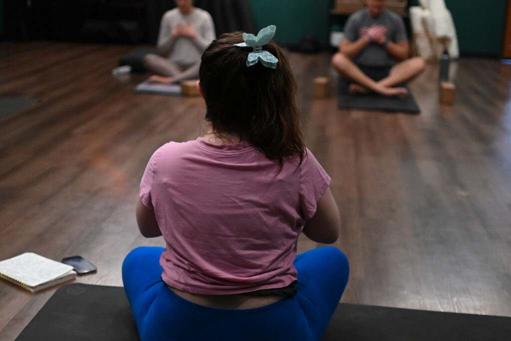 Anastasia Renfro, a white woman with long brown hair wearing a pink crop top , blue leggings and a light blue butterfly clip leads a class through meditation.