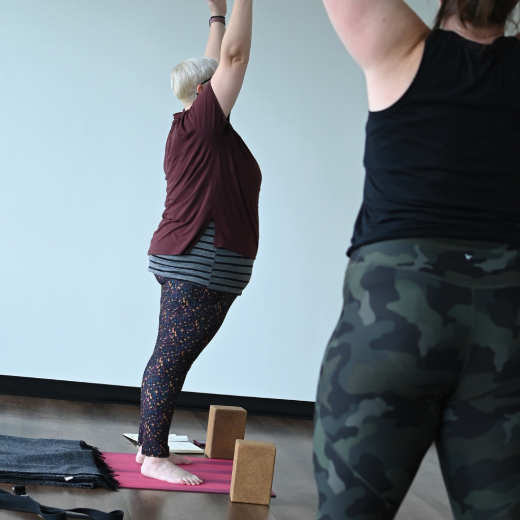 Karen Kirchman, w hite woman with platinum blonde hair wearing a dark maroon shirt with black leggings leads a yoga class.