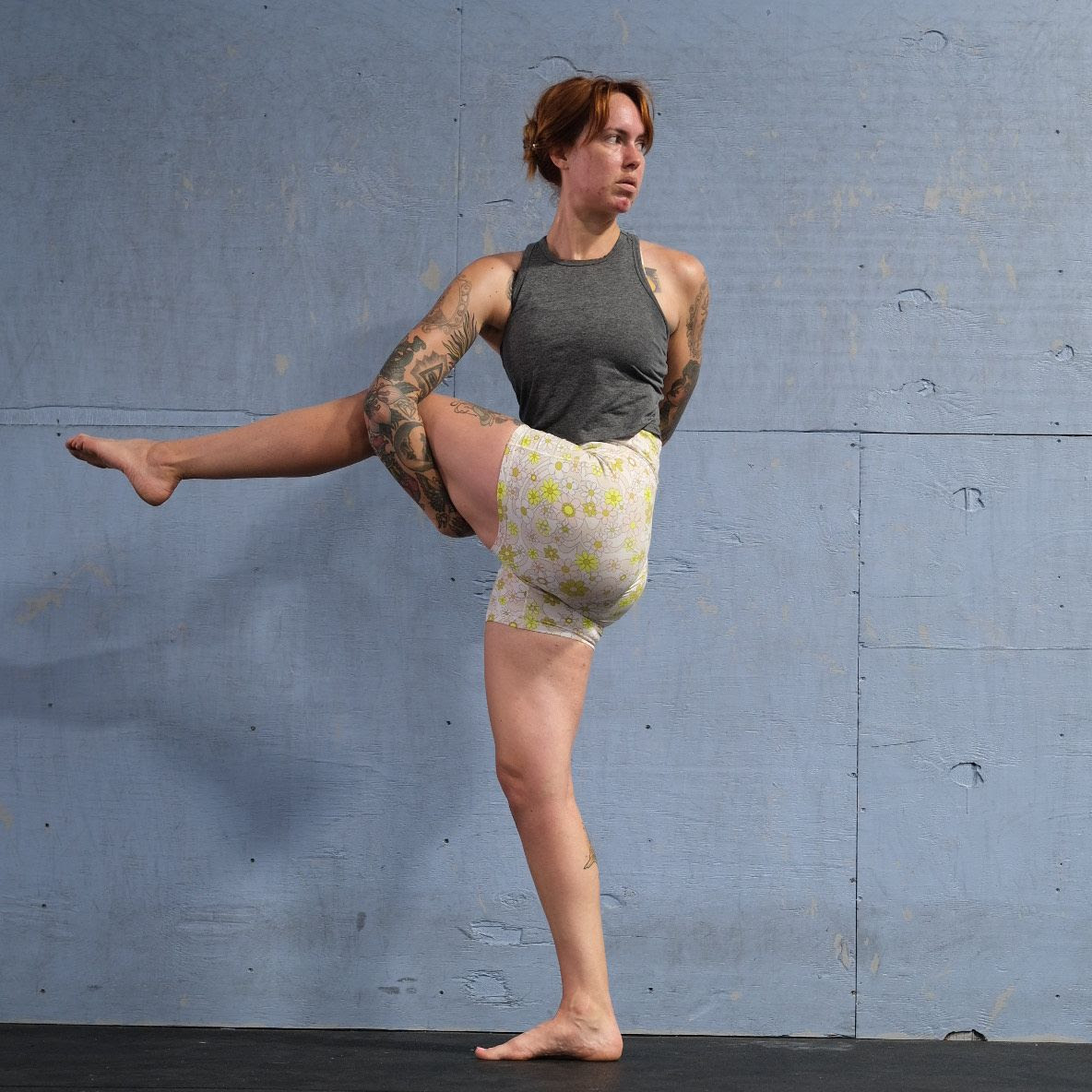 Marta Gruber, a white woman with red hair wearing white yoga shorts and a grey tank top practices  Parvrita Hasta Padanghtustasana (dancing Shiva)