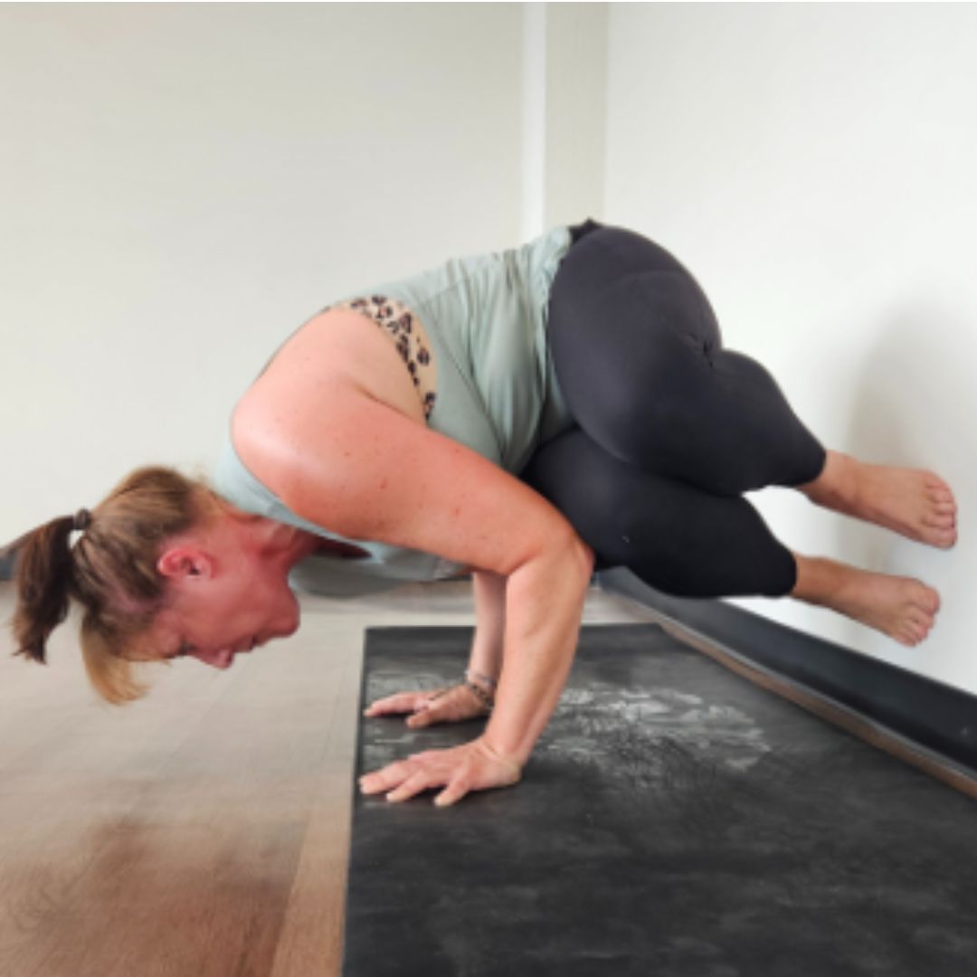 Kati Black, a white woman with ight brown hair wearing black leggings and a light blue tank top practices side crow pose supported at the wall.