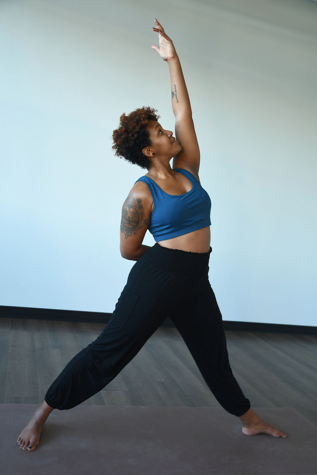 Arie McKenzie, a black woman with short dark hair, wearing a blue crop top and black pants practices sky archer pose on a beige yoga mat. 