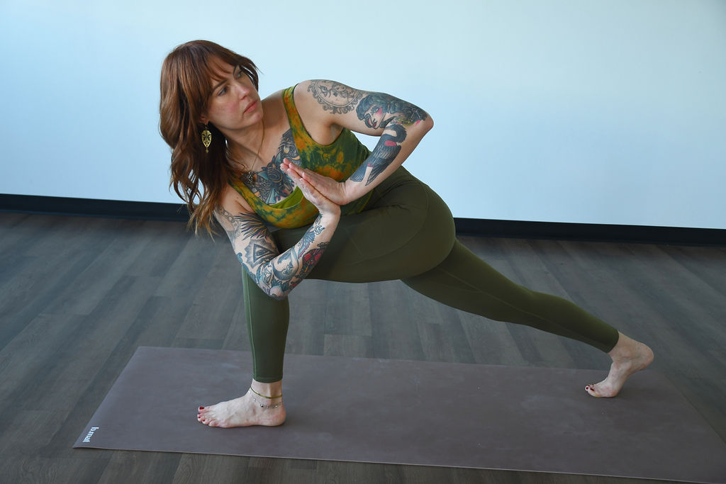 Marta Gruber, a white woman with red hair wearing green leggings and green crop to practices prayer twist lunge on a beige mat. 