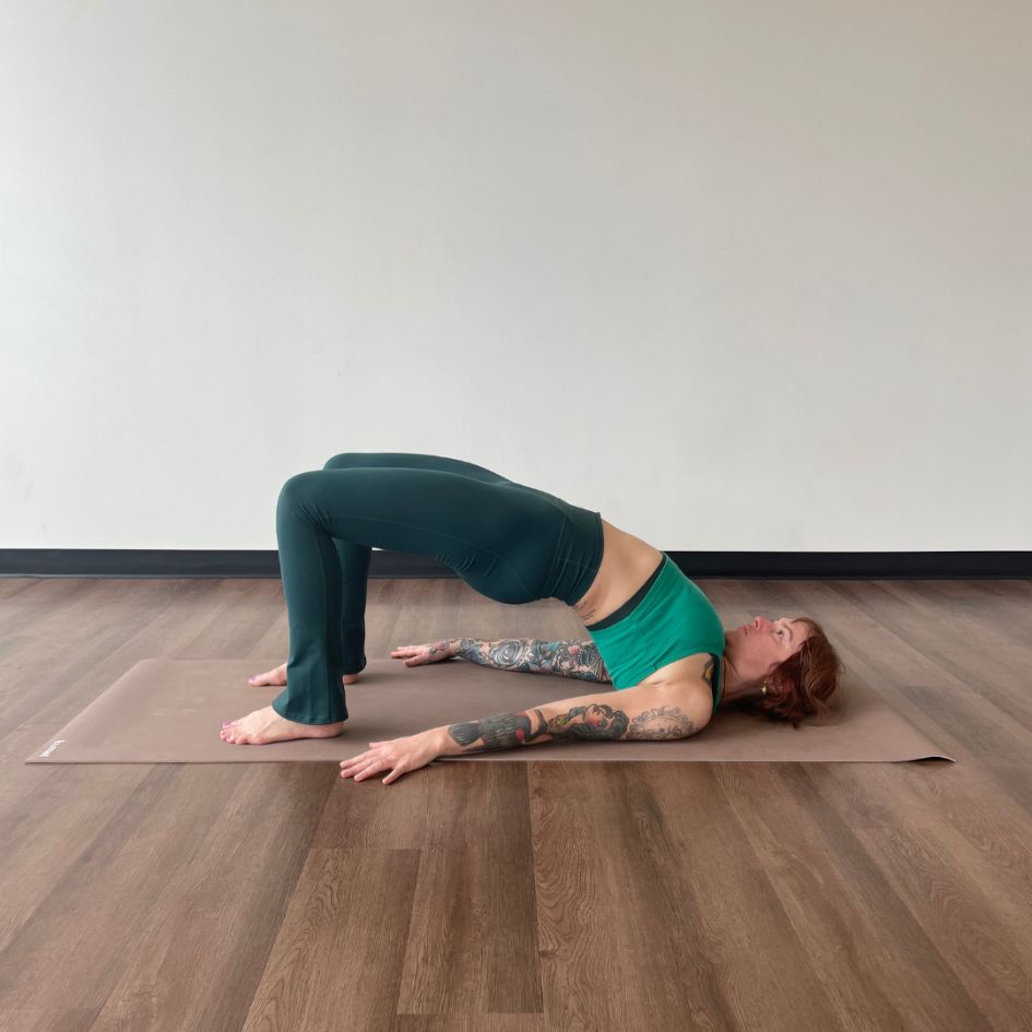Marta Gruber, a white woman with red hair wearing a bright green crop top and dark green leggings practices bridge pose on a beige yoga mat. 