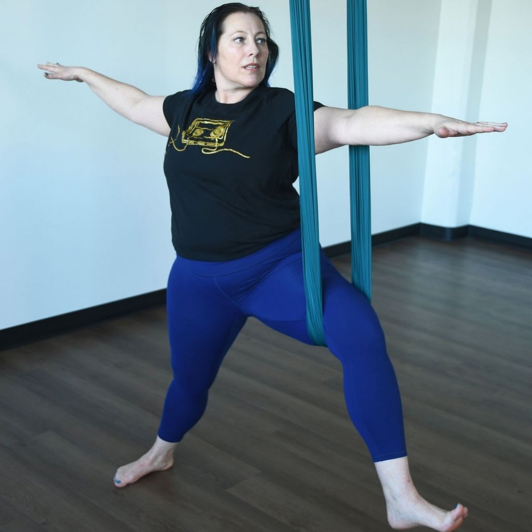 Kati Black , a white woman with dark blue hair wearing blue leggings and a black shirt practices warrior two with an aerial silk.