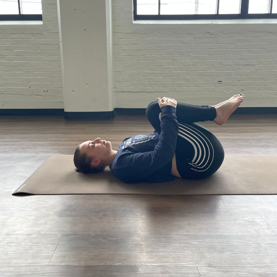 Zoe Lowden, a white woman with brown hair wearing a navy blue sweatshirt and black leggings with white stripes practices apanasana on a beige yoga mat. 