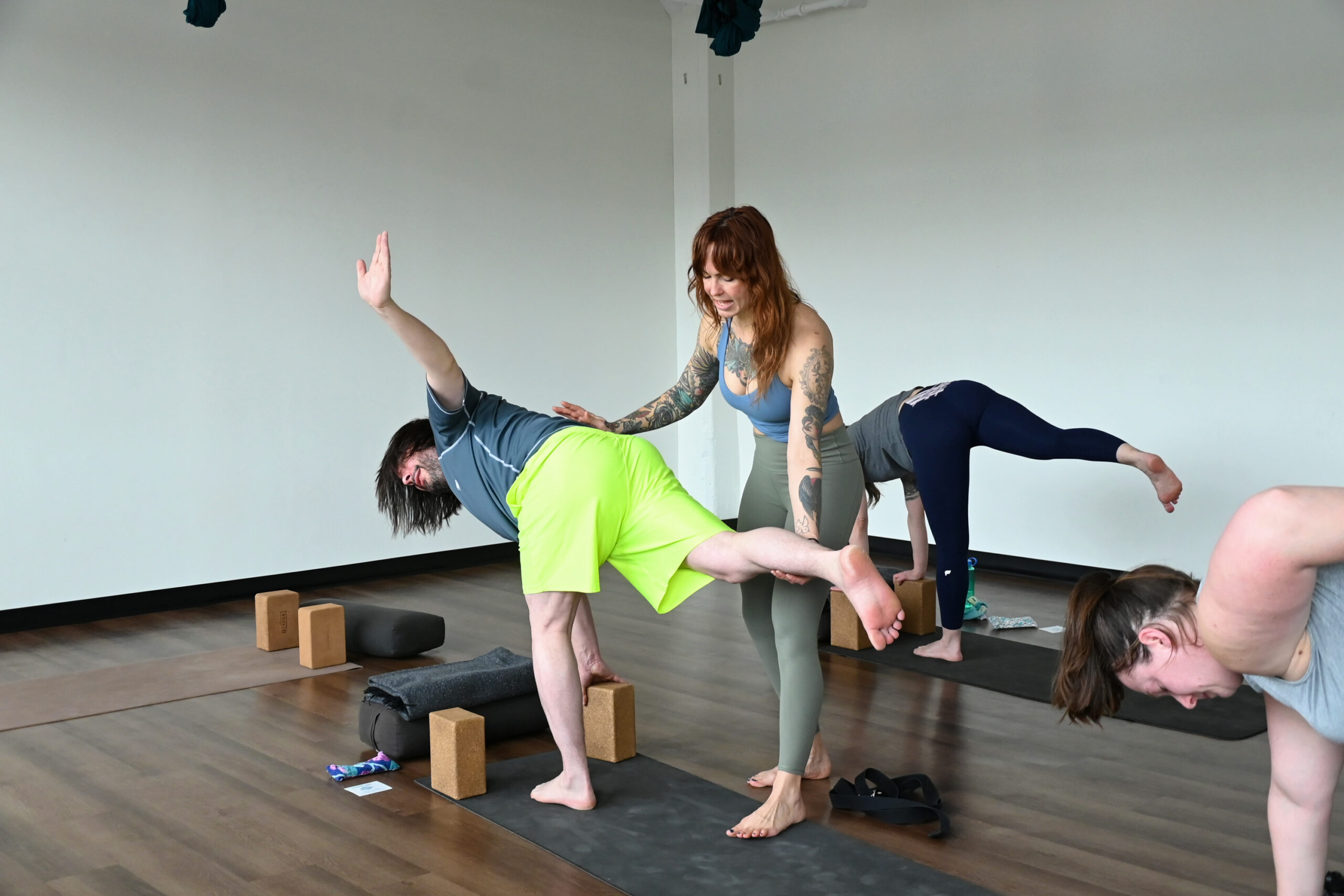 Marta Gruber, a white woman with red hair wearing a grey tank top and grey leggings assists a male student  wearing green shorts and a grey shirt practicing ardha chandrasana. 