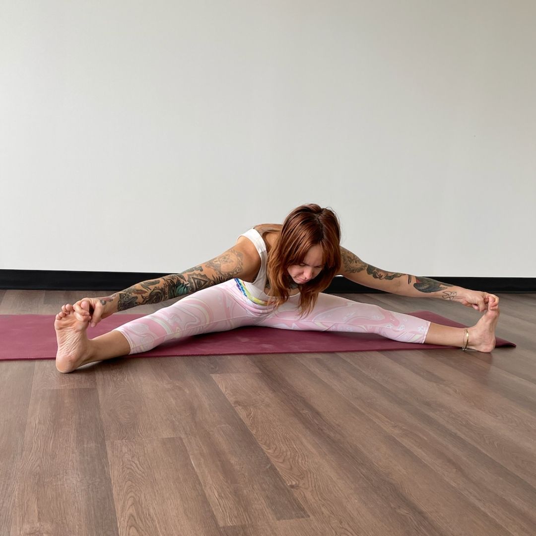 Marta Gruber, a white woman with red hair wearing light pink leggings and a white tank top sits in upavhista konasana on a dark red yoga mat. 