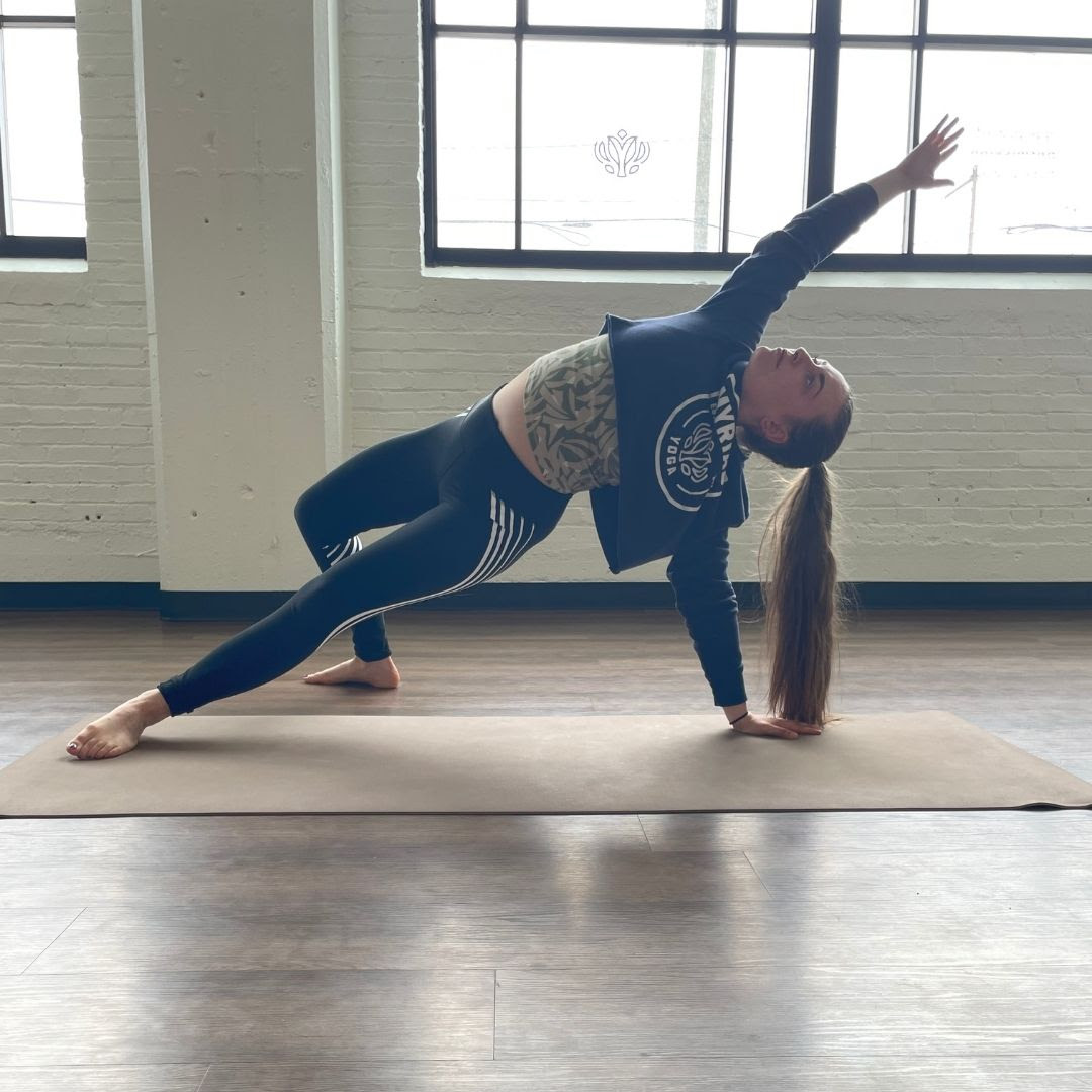 Zoe Lowden, a white woman with brown hair wearing black leggings and a blue sweatshirt practices camatakarasana on a beige yoga mat in front of brightly lit windows. 