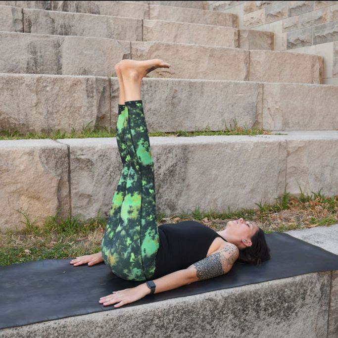 Jordan Nommay, a white woman with brown hair wearing green leggins and a black tank top practices legs up the wall on a black yoga mat outside. 
