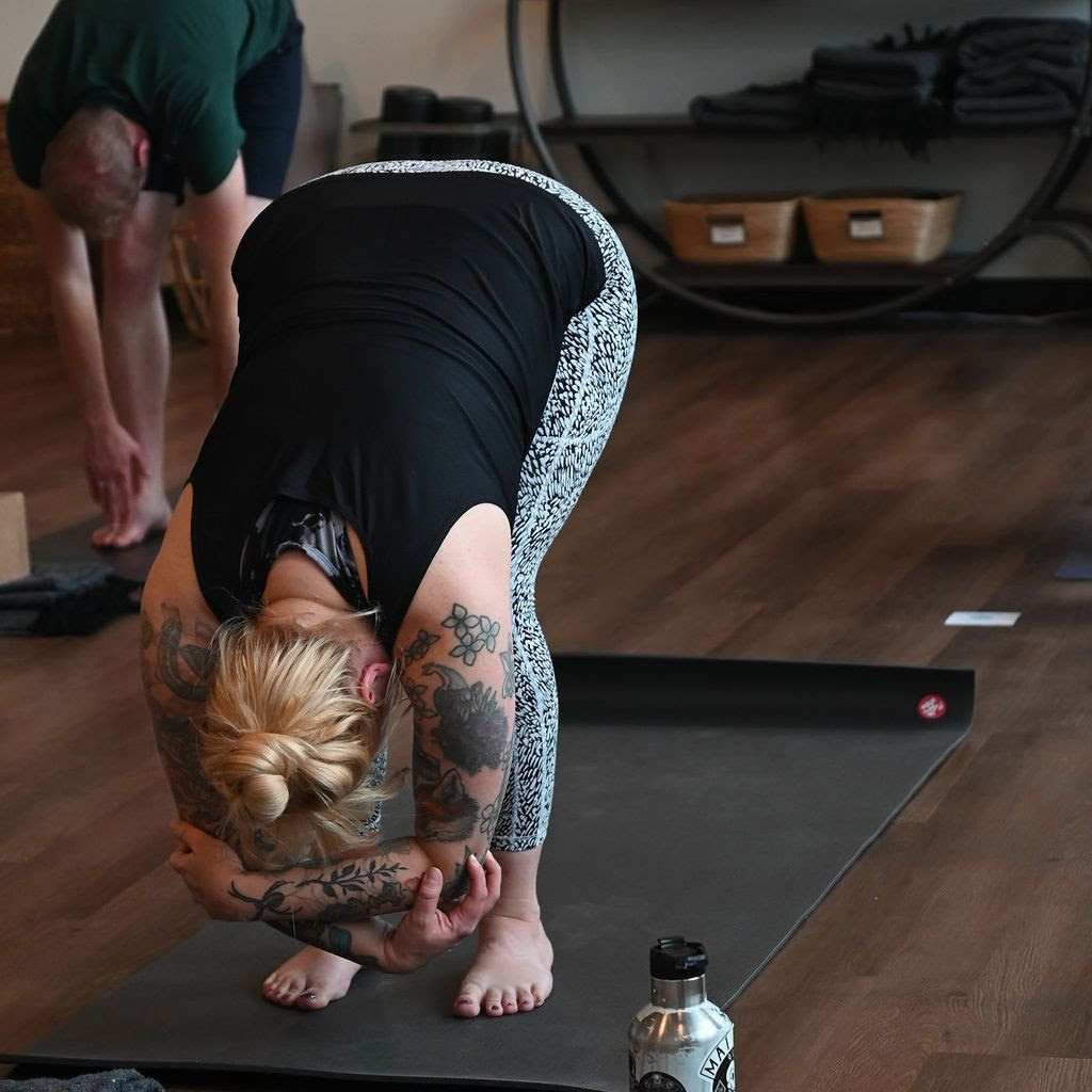Lauren Patton, a white woman with blonde hair pulled into a bun wearing patterned leggings and a black tank top practices uttanasana on a black yoga mat during a yoga class. 